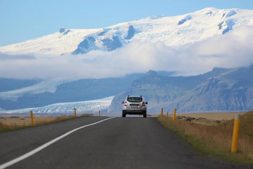 Scenic highway photograph showing a white SUV driving on a paved road through dramatic Icelandic landscape. Snow-capped mountains and glaciers are visible in the background under a partly cloudy sky. The road has yellow reflector posts along the sides, and the terrain features brown grassland. The image captures the vastness and beauty of remote driving conditions. #highway #SUV